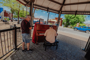 Deux hommes autour du piano public à la place de la Seigneurie. Un homme joue du piano. Le second écoute.