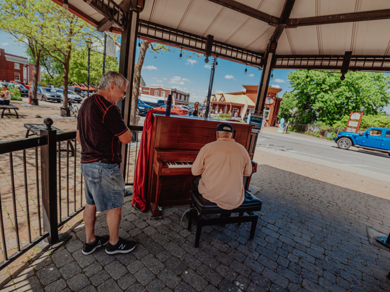 Deux hommes autour du piano public à la place de la Seigneurie. Un homme joue du piano. Le second écoute.
